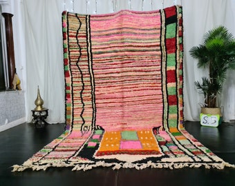May include: A large, colorful Moroccan rug with a pink, red, and green striped pattern. The rug has a black border with geometric designs. The rug is laid out on a black floor.
