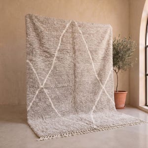 May include: A large, rectangular, shaggy area rug in a neutral taupe colour featuring a white diamond-shaped geometric pattern. The rug has a thick, textured pile and is finished with short, knotted fringe along the bottom edge of the fabric.