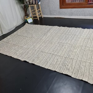 Puede incluir: Una alfombra rayada blanca y gris con textura tejida. La alfombra tiene un borde de flecos.