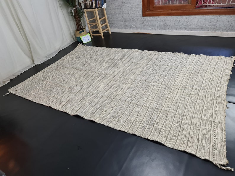 Puede incluir: Una alfombra rayada blanca y gris con textura tejida. La alfombra tiene un borde de flecos.