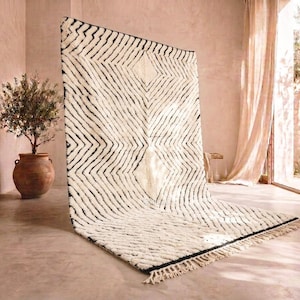 Puede incluir: Una alfombra grande de color crema con un patrón geométrico negro. La alfombra tiene un diseño en forma de diamante en el centro y una franja a lo largo del borde inferior. La alfombra se exhibe en una habitación con una paleta de colores neutros y luz natural.