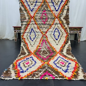 May include: A white, brown, blue, pink, and yellow Moroccan rug with a diamond pattern. The rug is handwoven and has a fringe on the ends.