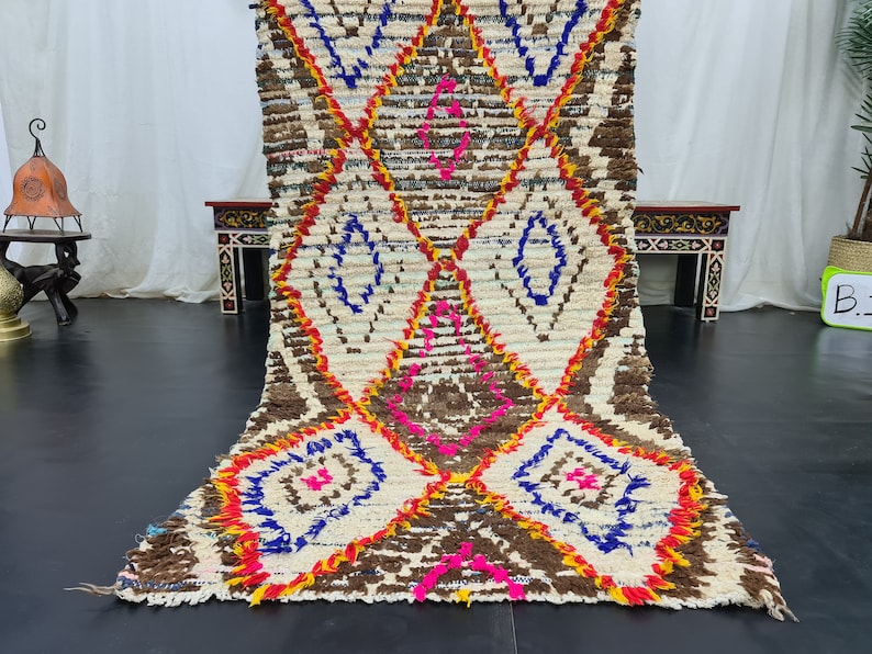 May include: A white, brown, blue, pink, and yellow Moroccan rug with a diamond pattern. The rug is handwoven and has a fringe on the ends.