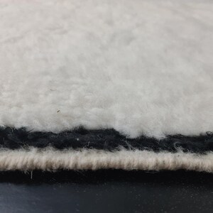 May include: Close-up of the edge of a white fluffy rug with a black trim. The rug has a soft, plush texture.