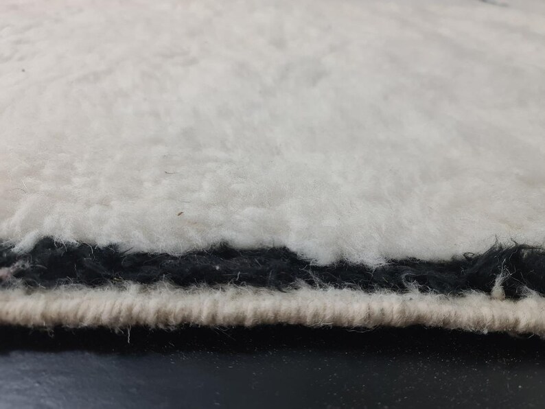 May include: Close-up of the edge of a white fluffy rug with a black trim. The rug has a soft, plush texture.