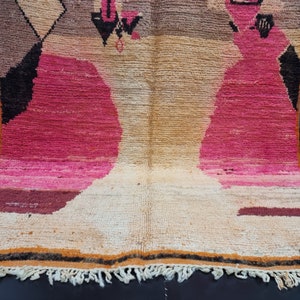 May include: A hand-woven rug with a geometric pattern in shades of pink, brown, and beige. The rug has a fringe border.