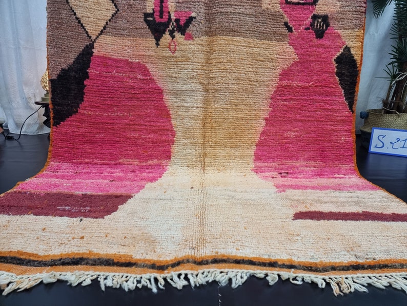 May include: A hand-woven rug with a geometric pattern in shades of pink, brown, and beige. The rug has a fringe border.