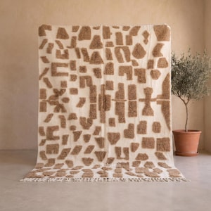 Puede incluir: Una alfombra rectangular con una base de color crema y un patrón repetitivo de formas geométricas en relieve, peludas y de color marrón claro. La alfombra tiene un pelo texturizado e incluye un fleco corto en el borde inferior.
