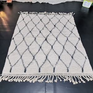 May include: A white and black Moroccan rug with a diamond pattern and fringe. The rug is made of wool and has a soft, fluffy texture.