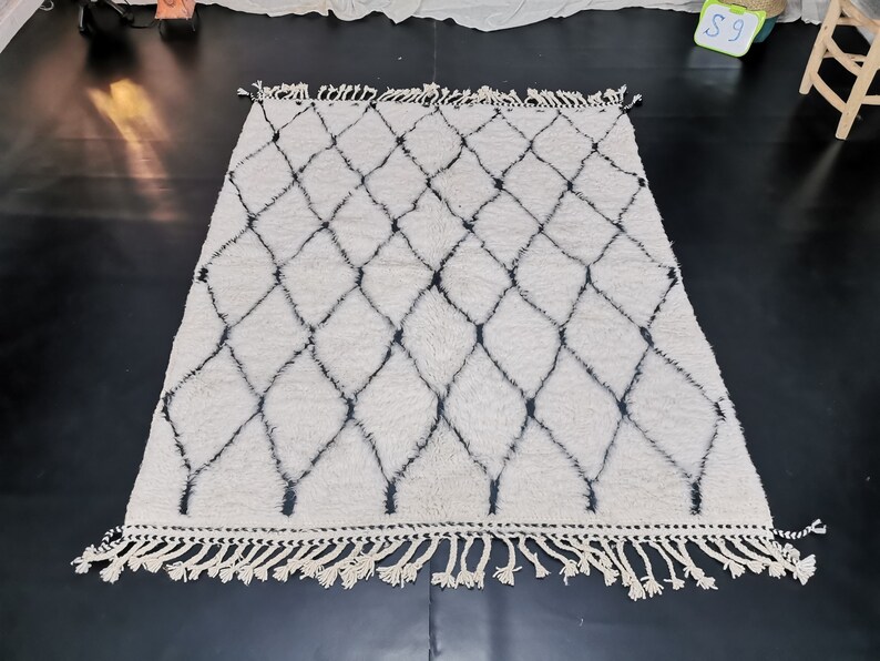 May include: A white and black Moroccan rug with a diamond pattern and fringe. The rug is made of wool and has a soft, fluffy texture.