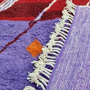 May include: A purple and red Moroccan rug with white fringe. The rug has a geometric pattern with orange and turquoise accents.