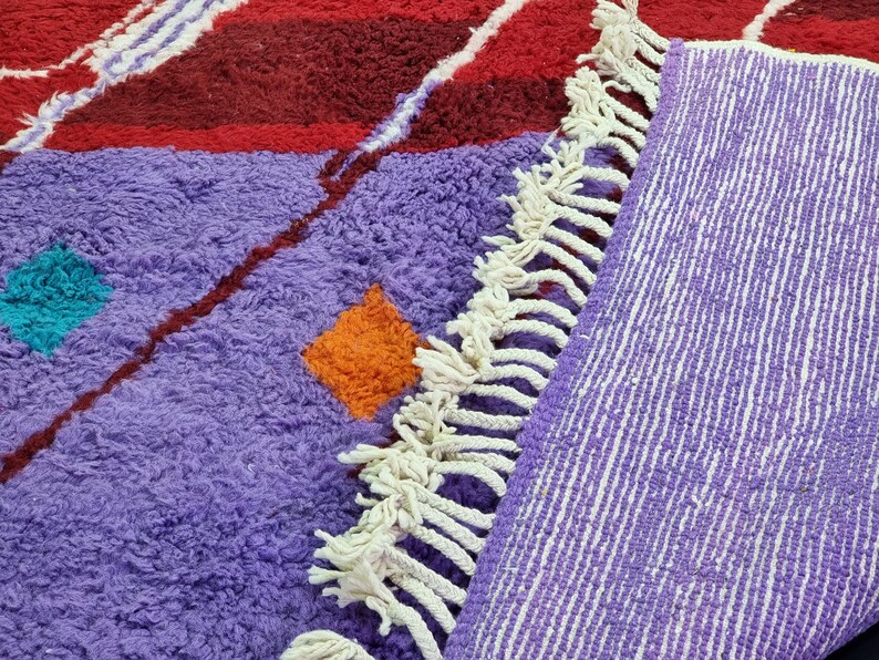 May include: A purple and red Moroccan rug with white fringe. The rug has a geometric pattern with orange and turquoise accents.