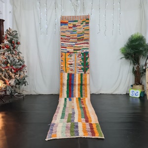 Puede incluir: Una alfombra marroquí larga y colorida con patrones geométricos en tonos de rojo, naranja, amarillo, verde, azul y blanco. La alfombra está colocada sobre un suelo de madera en una habitación con un árbol de Navidad y una planta.