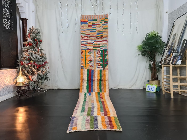 May include: A long, colorful Moroccan rug with geometric patterns in shades of red, orange, yellow, green, blue, and white. The rug is laid out on a wooden floor in a room with a Christmas tree and a plant.