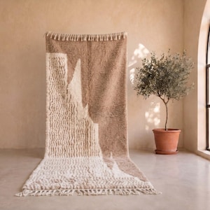 Peut inclure: Un tapis rectangulaire à poils longs présentant un design bicolore en beige et crème. Le tapis affiche un motif géométrique abstrait avec des franges sur les bords supérieur et inférieur, mettant en valeur une surface douce et touffue.