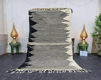 May include: Black and white striped rug with a fringe border. The rug is made of wool and has a geometric pattern. The rug is displayed on a gray floor with two gold lamps and side tables.