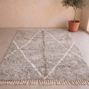 May include: A rectangular area rug featuring a thick, shaggy pile in a mottled grey colour. The rug is decorated with a large, white, hand-drawn diamond pattern that spans the centre. The short ends of the rug are finished with cream-coloured fringe.