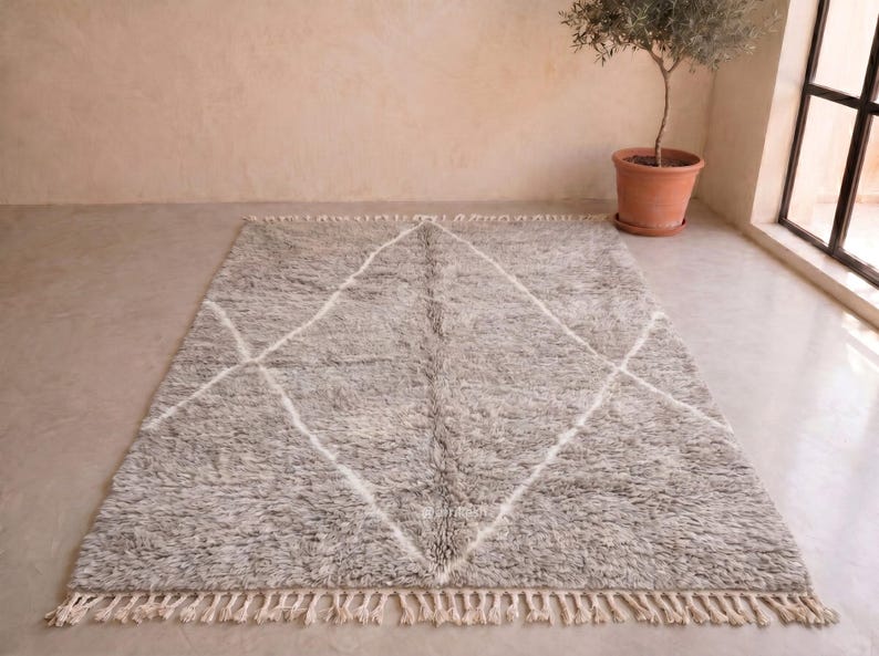 May include: A rectangular area rug featuring a thick, shaggy pile in a mottled grey colour. The rug is decorated with a large, white, hand-drawn diamond pattern that spans the centre. The short ends of the rug are finished with cream-coloured fringe.