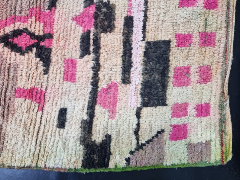 May include: A close-up of a colorful woven rug with pink, black, and white geometric patterns. The rug has a frayed edge and a textured surface.
