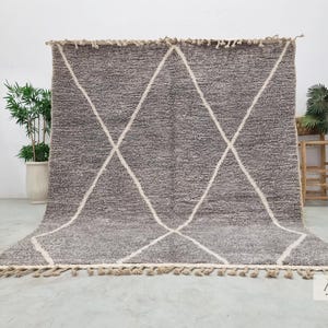 Puede incluir: Una alfombra marroquí gris y blanca con un patrón de diamantes. La alfombra tiene una superficie texturizada y una franja a lo largo del borde inferior. La alfombra se exhibe sobre un suelo de hormigón.