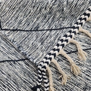 May include: Black and white geometric patterned rug with fringe. The rug has a woven texture and a black and white striped fringe.