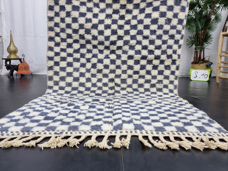 Op de afbeelding: Een wit en blauw geruit tapijt met een franje rand. Het tapijt is gemaakt van wol en heeft een zachte, pluche textuur.