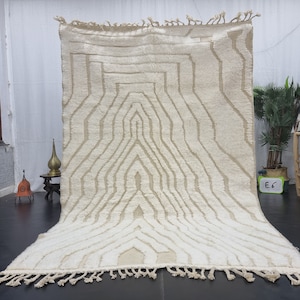 May include: A white Moroccan rug with a geometric pattern. The rug has a fringe border and is made of wool.