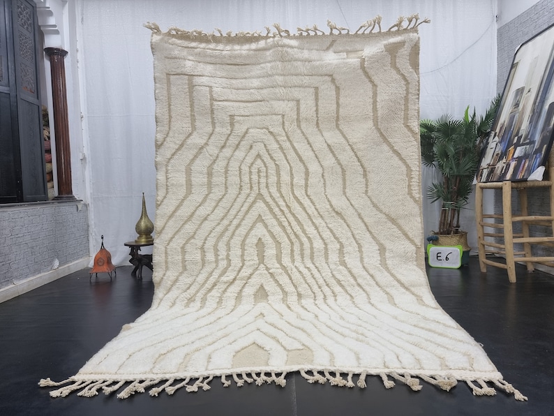 May include: A white Moroccan rug with a geometric pattern. The rug has a fringe border and is made of wool.