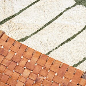 May include: A woven leather chair with a light brown hue is positioned diagonally, partially obscuring a textured rug. The rug features a cream-colored base with dark green stripes. The chair's design showcases a grid-like pattern.