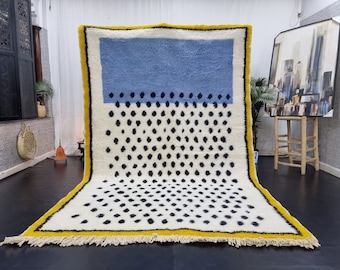 May include: A white wool rug with a blue and black geometric pattern. The rug has a yellow border and fringe.