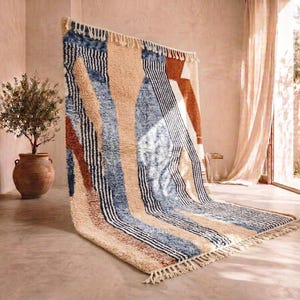 Könnte beinhalten: Ein großer, rechteckiger Teppich mit geometrischem Design. Der Teppich hat vertikale Streifen in Beige, Blau und Braun mit einem weißen Fransen am unteren Rand. Der Teppich wird in Innenräumen ausgestellt.