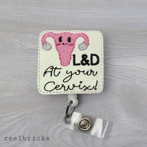 May include: White glitter badge holder with a pink embroidered uterus with a smiling face and the text "L&D At your Cervix!"