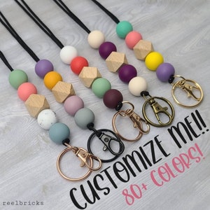 May include: A variety of colorful beaded lanyards with different colored beads and wooden beads. Each lanyard has a metal clasp. The text "Customize Me! 80+ Colors!" is written on the image.