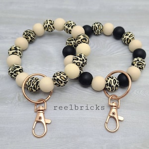 May include: Two beige and black beaded keychains with gold-tone lobster clasps and key rings. The beads have a leopard print pattern. The keychains are labeled "reelbricks".