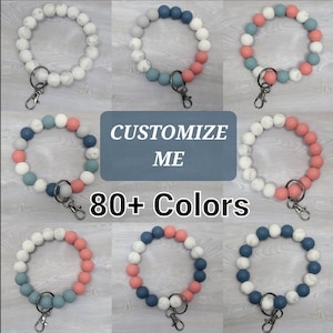 May include: Assortment of colorful beaded keychains with a silver key ring. The beads are in various colors, including white, blue, and pink. The keychains are arranged in a grid pattern. The text "CUSTOMIZE ME" and "80+ Colors" is displayed on the image.