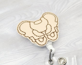 May include: A white and brown embroidered badge reel with a design of a human pelvis. The reel is attached to a clear plastic badge holder with a metal clip.
