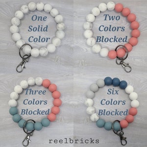 May include: Four silicone beaded keychains in different color combinations. The first keychain is all white with a marble pattern. The second keychain is white with marble pattern and pink. The third keychain is white with marble pattern, pink, and blue. The fourth keychain is gray, blue, and pink. All keychains have a silver key ring.