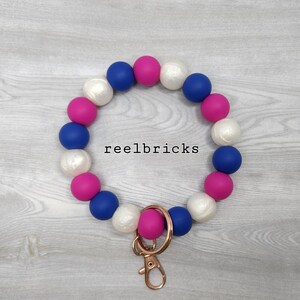 May include: A pink, blue, and white beaded keychain with a gold metal key ring. The beads are arranged in a repeating pattern. The keychain is on a wooden surface. reelbricks