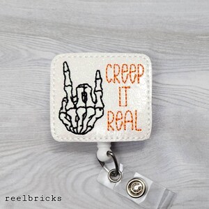 May include: White glitter badge holder with a black skeleton hand making the rock and roll sign. The text "Creep It Real" is in orange.