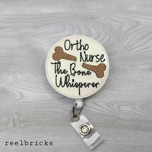 May include: White round badge holder with a white background and brown embroidery. The text reads "Ortho Nurse, The Bone Whisperer" with two brown bone images.