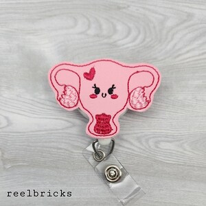 May include: Pink felt anatomical heart with a smiling face and a small red heart above it. The heart is attached to a clear plastic retractable badge holder.