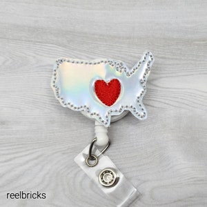 May include: A silver iridescent USA map with a red heart in the center, attached to a white retractable badge holder.