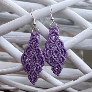 Puede incluir: Un par de pendientes de macramé morados hechos a mano. Los pendientes presentan un diseño en forma de diamante con intrincados nudos, suspendidos de ganchos plateados. Se exhiben sobre un fondo blanco tejido.