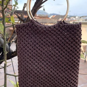Pu&ograve; includere: Una borsa tote in macram&egrave; marrone con manico in metallo dorato. La borsa &egrave; rettangolare con una trama intrecciata. Il manico ha un design circolare. La borsa &egrave; appesa a un ramo d'albero.