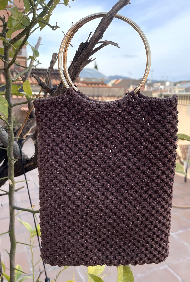 Pu&ograve; includere: Una borsa tote in macram&egrave; marrone con manico in metallo dorato. La borsa &egrave; rettangolare con una trama intrecciata. Il manico ha un design circolare. La borsa &egrave; appesa a un ramo d'albero.