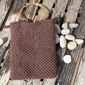 Pu&ograve; includere: Una borsa tote marrone all'uncinetto con un manico circolare color oro. La borsa &egrave; rettangolare e ha una superficie strutturata. La borsa &egrave; su una superficie di legno stagionato con diverse pietre lisce e chiare.
