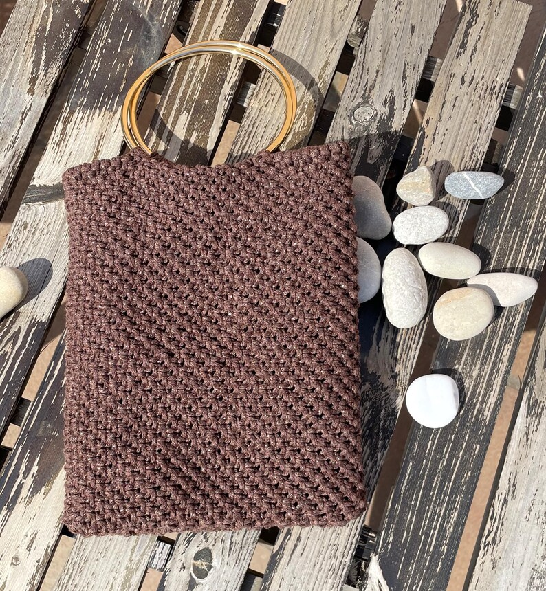 Pu&ograve; includere: Una borsa tote marrone all'uncinetto con un manico circolare color oro. La borsa &egrave; rettangolare e ha una superficie strutturata. La borsa &egrave; su una superficie di legno stagionato con diverse pietre lisce e chiare.