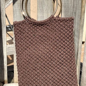 Pu&ograve; includere: Una borsa tote in macram&egrave; marrone con manici ad anello in metallo dorato. La borsa ha una forma rettangolare e una trama intrecciata. I manici sono circolari e attaccati alla parte superiore della borsa.