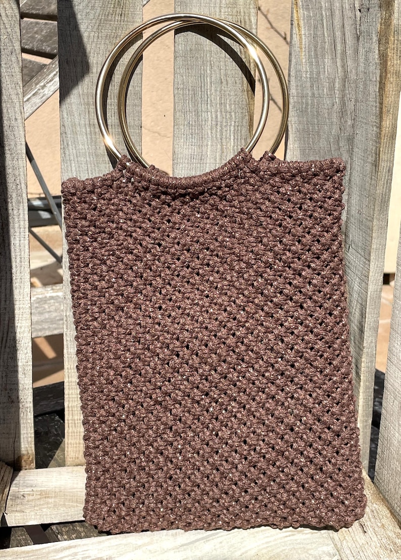 Pu&ograve; includere: Una borsa tote in macram&egrave; marrone con manici ad anello in metallo dorato. La borsa ha una forma rettangolare e una trama intrecciata. I manici sono circolari e attaccati alla parte superiore della borsa.