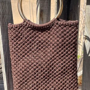 Pu&ograve; includere: Una borsa tote in macram&egrave; marrone con manici in metallo dorato. La borsa &egrave; rettangolare con una trama intrecciata. Le maniglie circolari sono attaccate alla parte superiore. La borsa &egrave; appesa a una staccionata di legno.